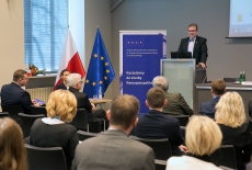 Dyrektor KSAP Wojciech Federczyk przemawia z mównicy w auli KSAP. Widoczni siedzący uczestnicy konferencji. Obok mównicy baner KSAP i flagi polska i unijna.