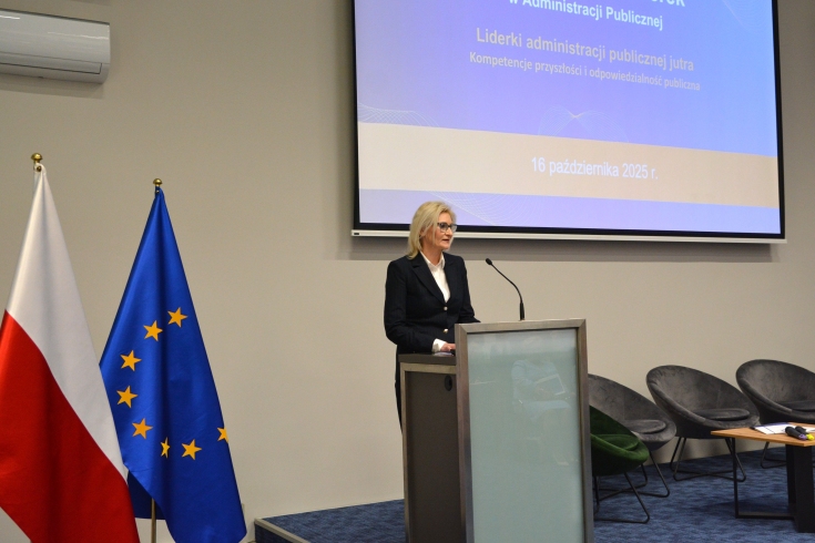 Przy mównicy stoi kobieta i przemawia.